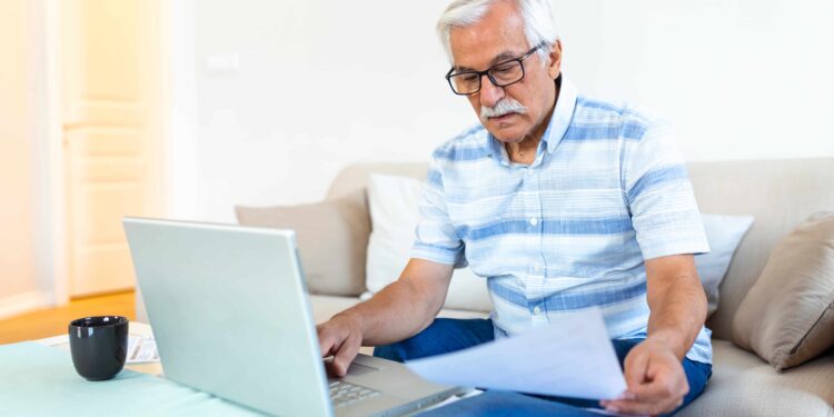 Idoso sentado no sofá usando notebook enquanto confere documentos