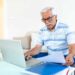 Idoso sentado no sofá usando notebook enquanto confere documentos