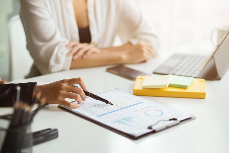 Análise de relatórios financeiros em reunião empresarial 2026. Duas pessoas analisam gráfico e tabela em relatório impresso sobre mesa de escritório.