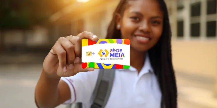 Jovem sorridente mostra cartão colorido do programa Pé de Meia em frente à escola.