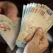 Pessoa contando várias cédulas de cinquenta reais com as mãos em ambiente interno.