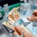 Pessoa segurando várias notas de real com cédulas de R$ 100, R$ 50 e R$ 20 frente a caixa eletrônico.