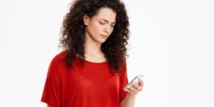 Mulher de camiseta vermelha olha pensativa para celular em ambiente claro