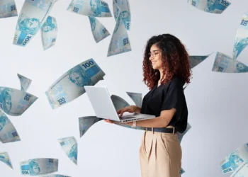 Mulher sorrindo digita em notebook, cercada por notas de R$ 100 em fundo claro.