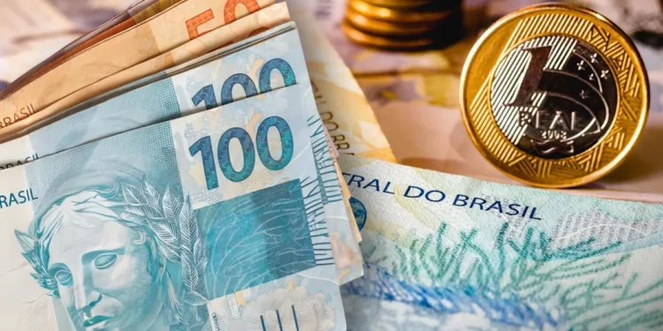 Cédulas de 100 e 50 reais agrupadas com moedas douradas ao fundo sobre mesa.
