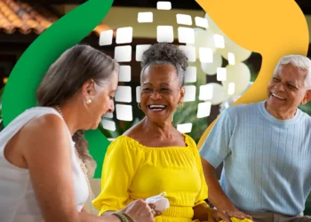 Três idosos felizes sentados juntos, um deles em destaque sorrindo e usando blusa amarela, ambiente descontraído