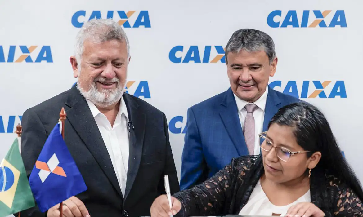 Autoridades da Caixa formalizam concessão de crédito habitacional 2026. Três pessoas em evento da Caixa Econômica, mulher assinando documento com autoridades ao fundo.