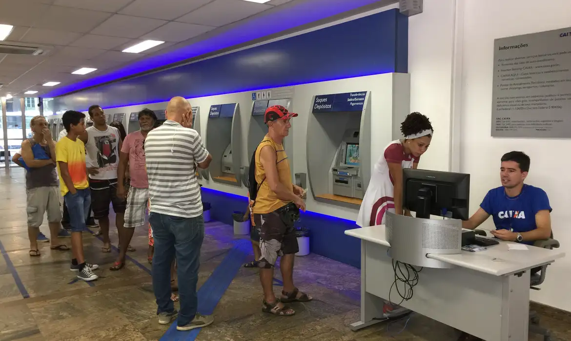 Clientes utilizando caixas eletrônicos e atendimento bancário em 2026. Pessoas em fila usando caixas eletrônicos enquanto atendente auxilia cliente em computador.