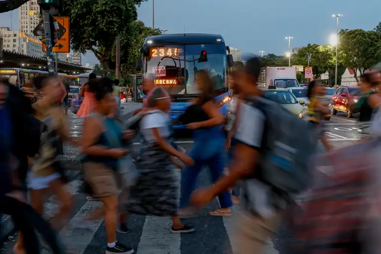 Pedestres atravessando avenida movimentada no centro urbano Pessoas atravessam faixa de pedestres em frente a ônibus azul em avenida movimentada ao entardecer