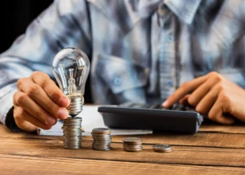 Pessoa empilhando moedas e segurando lâmpada ao lado de calculadora sobre mesa de madeira