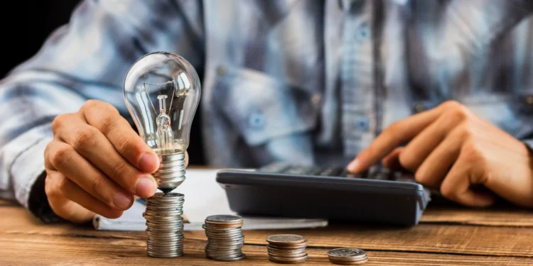 Pessoa empilhando moedas e segurando lâmpada ao lado de calculadora sobre mesa de madeira