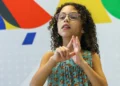 Menina com cabelos cacheados e óculos usando linguagem de sinais em frente a parede colorida, representando crianças com deficiência que seriam beneficiadas pelo Auxílio Mãe Atípica