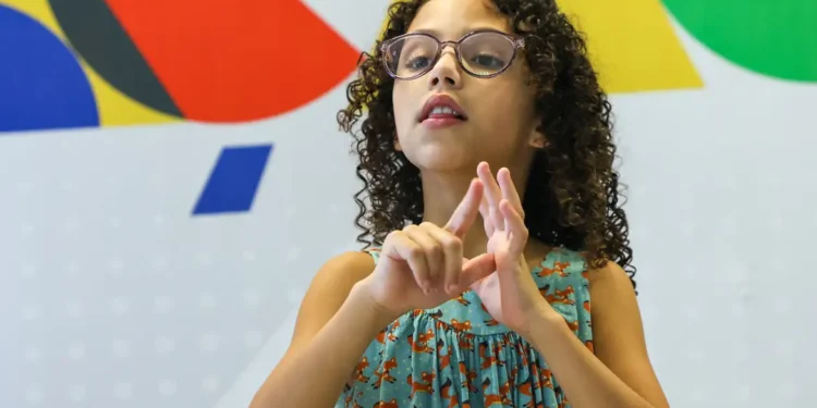 Menina com cabelos cacheados e óculos usando linguagem de sinais em frente a parede colorida, representando crianças com deficiência que seriam beneficiadas pelo Auxílio Mãe Atípica