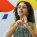 Menina com cabelos cacheados e óculos usando linguagem de sinais em frente a parede colorida, representando crianças com deficiência que seriam beneficiadas pelo Auxílio Mãe Atípica