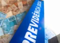 Placa azul vertical com a inscrição "PREVIDÊNCIA SOCIAL" em letras brancas. Em primeiro plano, notas de Real de 50 e 100 estão distribuídas de forma sobreposta. Ao fundo, a estrutura de um edifício moderno reflete a claridade externa.