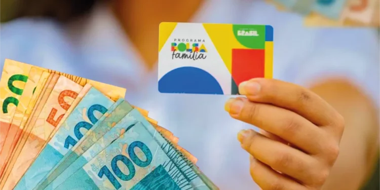 Mão segurando o cartão do Programa Bolsa Família ao lado de notas de R$ 100 e R$ 50, representando o pagamento do benefício em março de 2026