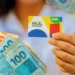 Mão segurando o cartão do Programa Bolsa Família ao lado de notas de R$ 100 e R$ 50, representando o pagamento do benefício em março de 2026