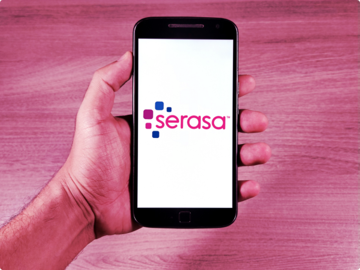 Mão segurando celular com logotipo da Serasa na tela sobre fundo rosa