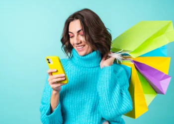 Mulher sorrindo segura várias sacolas de compras coloridas enquanto usa um celular amarelo