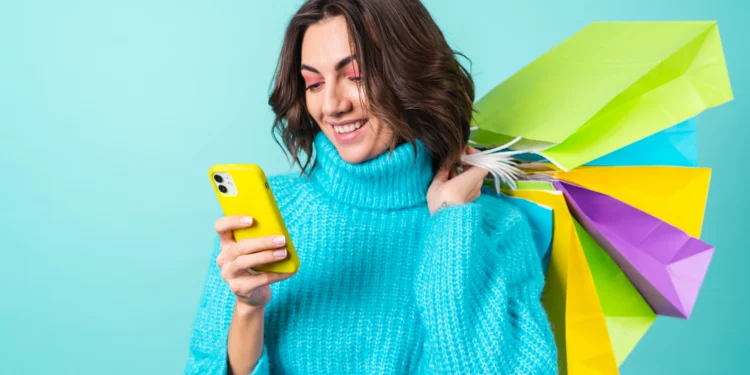 Mulher sorrindo segura várias sacolas de compras coloridas enquanto usa um celular amarelo
