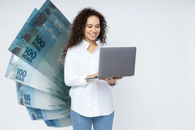 Jovem sorridente acessando notebook com notas de 100 reais ao fundo