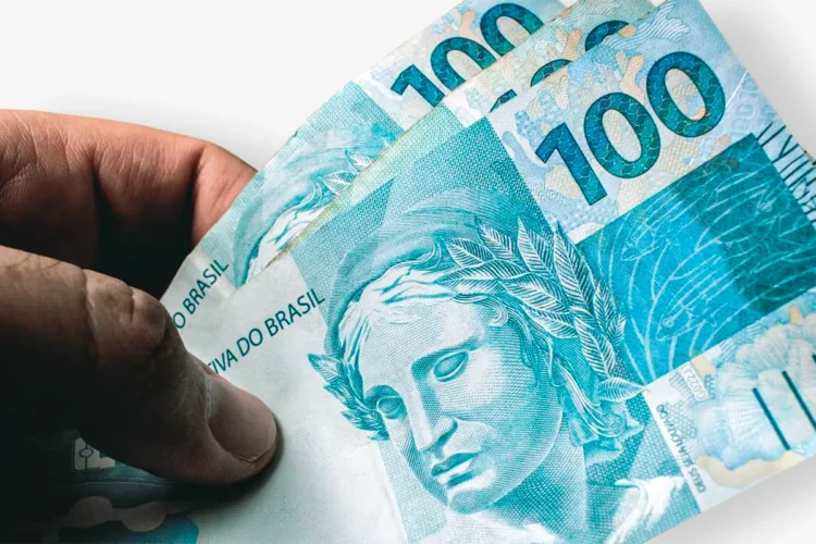 Mão conta notas de cem reais sobre fundo claro, destacando valor