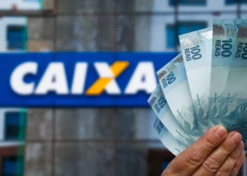 Mão segurando várias notas de 100 reais com letreiro da Caixa ao fundo