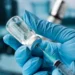 Mãos com luvas azuis seguram seringa extraindo líquido de frasco transparente