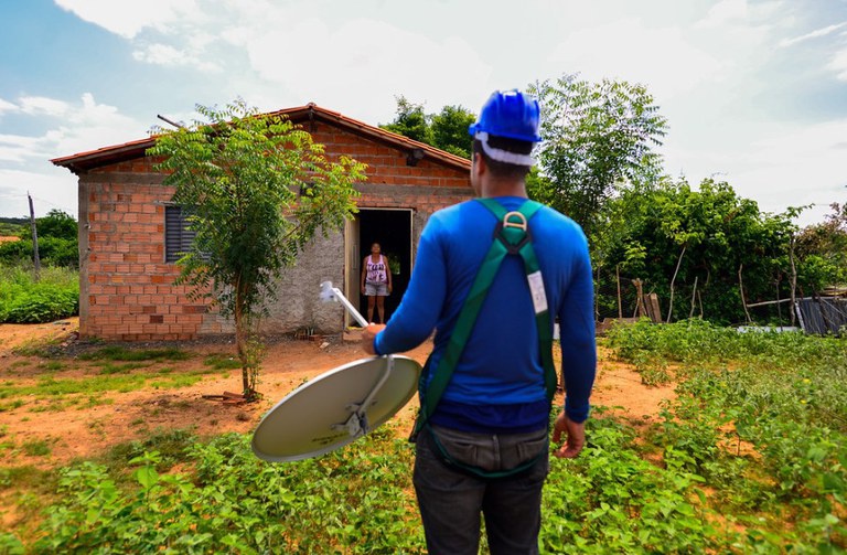 Técnico de capacete azul carrega antena parabólica digital em direção à casa de tijolos em área rural.
