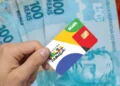 Close de uma mão segurando o cartão oficial do programa Bolsa Família, com cores amarelo, azul, verde e vermelho. Ao fundo, cédulas de 100 reais desfocadas.