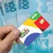 Close de uma mão segurando o cartão oficial do programa Bolsa Família, com cores amarelo, azul, verde e vermelho. Ao fundo, cédulas de 100 reais desfocadas.