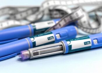 Três canetas injetáveis de cor azul, semelhantes às utilizadas para tratamento de diabetes e perda de peso, dispostas sobre uma superfície clara. Ao fundo, uma fita métrica cinza e branca está entrelaçada, simbolizando o monitoramento de medidas corporais.