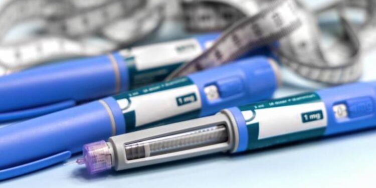 Três canetas injetáveis de cor azul, semelhantes às utilizadas para tratamento de diabetes e perda de peso, dispostas sobre uma superfície clara. Ao fundo, uma fita métrica cinza e branca está entrelaçada, simbolizando o monitoramento de medidas corporais.