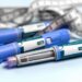Três canetas injetáveis de cor azul, semelhantes às utilizadas para tratamento de diabetes e perda de peso, dispostas sobre uma superfície clara. Ao fundo, uma fita métrica cinza e branca está entrelaçada, simbolizando o monitoramento de medidas corporais.