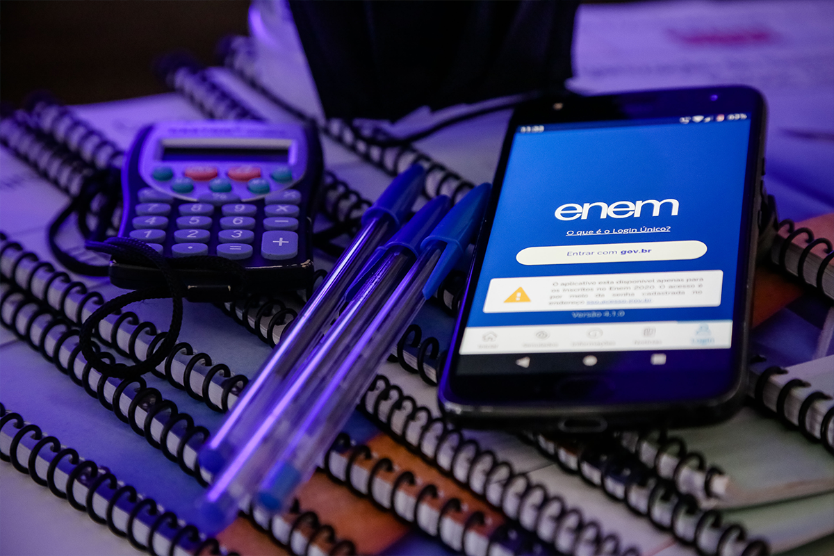 Um smartphone exibindo a tela de login do aplicativo Enem com o botão "Entrar com gov.br". O celular está sobre uma pilha de cadernos de espiral, ao lado de duas canetas azuis e uma calculadora pequena.