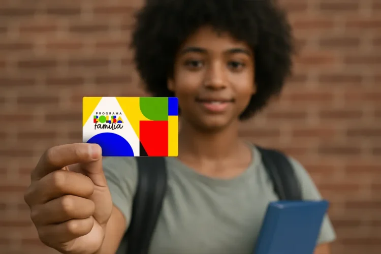 Jovem com cabelo cacheado segura cartão colorido do Bolsa Família e livro azul em frente a parede de tijolos