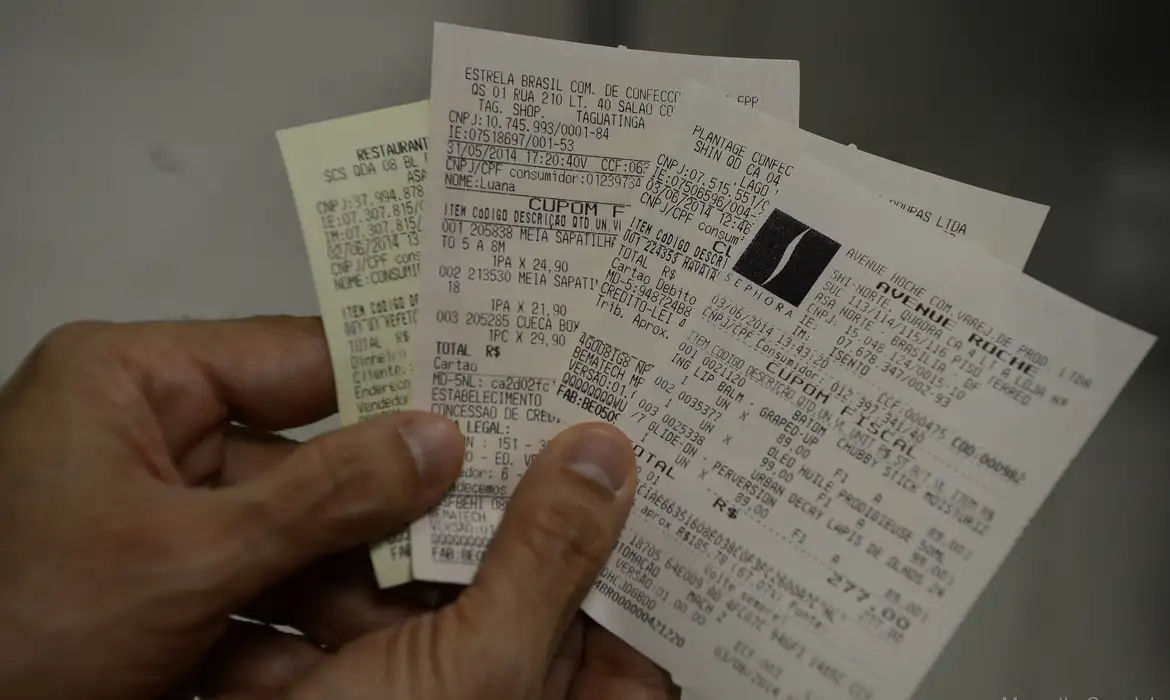 Mão segurando vários cupons fiscais, representando as notas com CPF que geram bilhetes para o sorteio da Nota Premiada Bahia