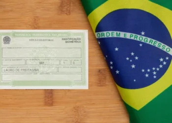 Título eleitoral da República Federativa do Brasil sobre mesa de madeira ao lado de bandeira nacional dobrada.