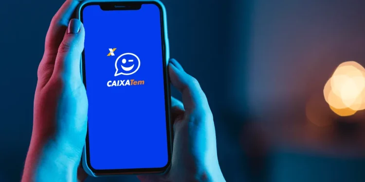 Mãos segurando celular com aplicativo Caixa Tem aberto na tela inicial azul com ícone sorridente.