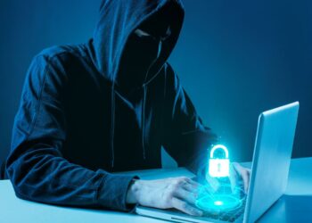 Ilustração de um hacker usando capuz e máscara escura em frente a um notebook em ambiente azulado. Sobre o teclado, há um holograma brilhante de um cadeado azul, simbolizando segurança digital e invasão de contas.