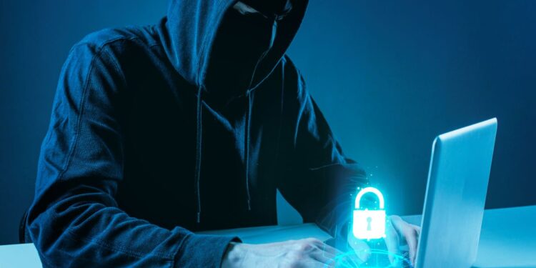 Ilustração de um hacker usando capuz e máscara escura em frente a um notebook em ambiente azulado. Sobre o teclado, há um holograma brilhante de um cadeado azul, simbolizando segurança digital e invasão de contas.