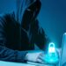 Ilustração de um hacker usando capuz e máscara escura em frente a um notebook em ambiente azulado. Sobre o teclado, há um holograma brilhante de um cadeado azul, simbolizando segurança digital e invasão de contas.