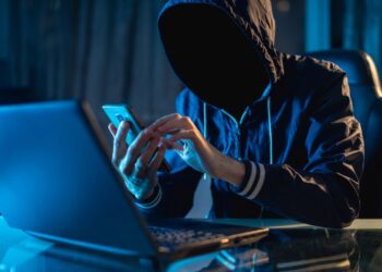 Um homem com capuz em um ambiente escuro, usando um celular e um notebook com iluminação azul, representando um hacker ou golpista digital em operação.