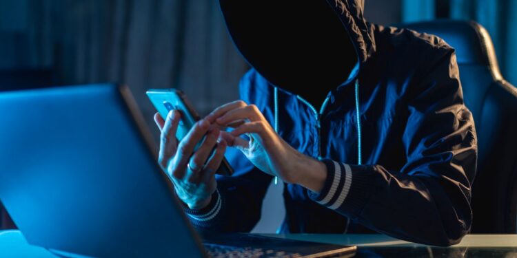 Um homem com capuz em um ambiente escuro, usando um celular e um notebook com iluminação azul, representando um hacker ou golpista digital em operação.