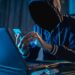 Um homem com capuz em um ambiente escuro, usando um celular e um notebook com iluminação azul, representando um hacker ou golpista digital em operação.