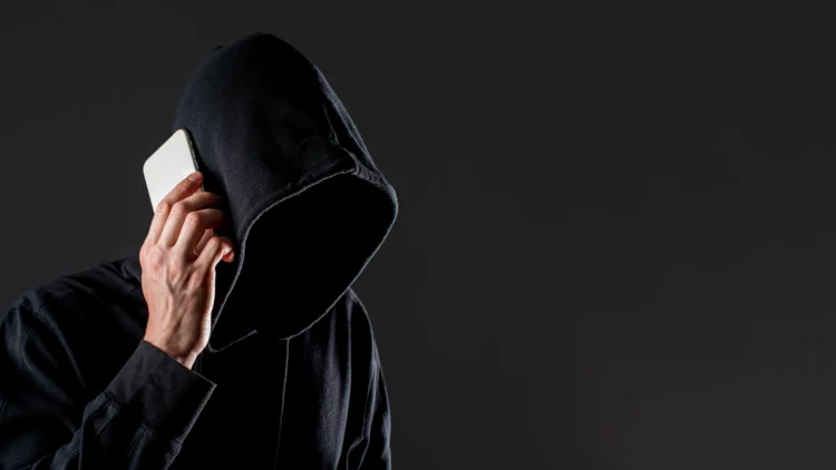Pessoa encapuzada de preto segura smartphone branco na orelha em fundo cinza escuro, representando golpe telefônico.