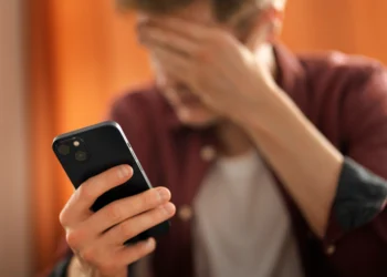 Jovem de camisa vinho segura smartphone preto enquanto cobre o rosto com a mão em sinal de frustração.