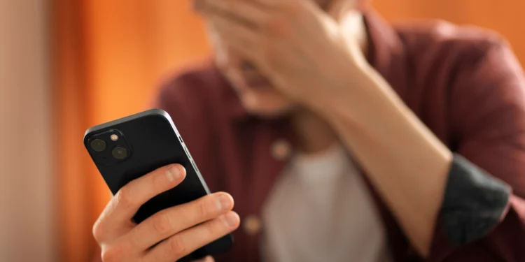 Jovem de camisa vinho segura smartphone preto enquanto cobre o rosto com a mão em sinal de frustração.