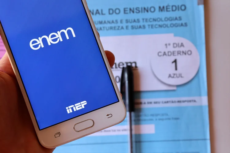 Mão segura celular com app Enem e logo INEP sobre caderno de provas azul do 1º dia com caneta preta.