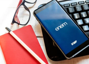 Celular mostrando tela azul do ENEM sobre teclado preto, ao lado de caderno vermelho, lápis e óculos.